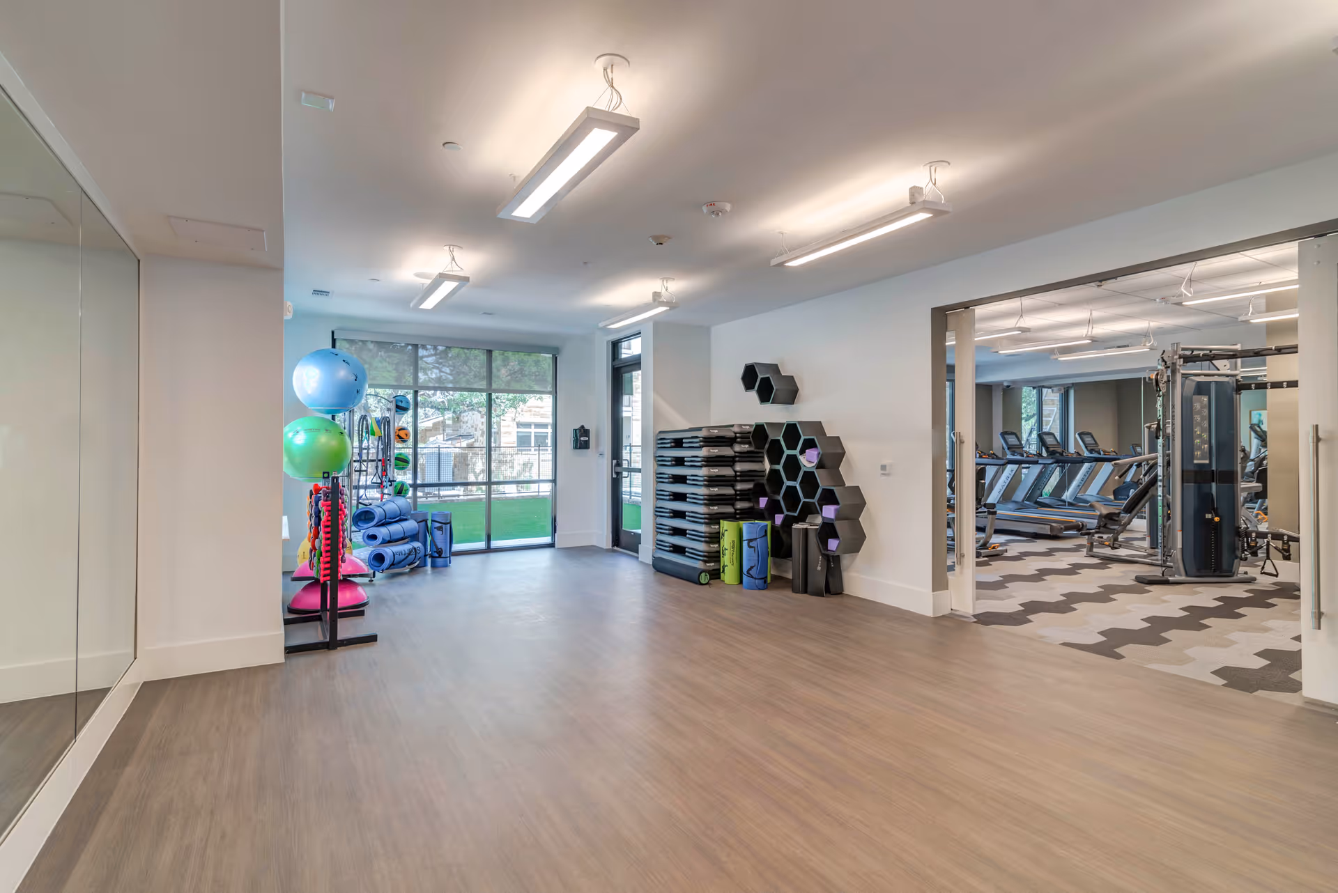 A spacious fitness room with exercise equipment including stability balls, yoga mats, and step platforms. The room has large windows and a door leading outside, with an adjacent room visible containing treadmills and other gym machines.