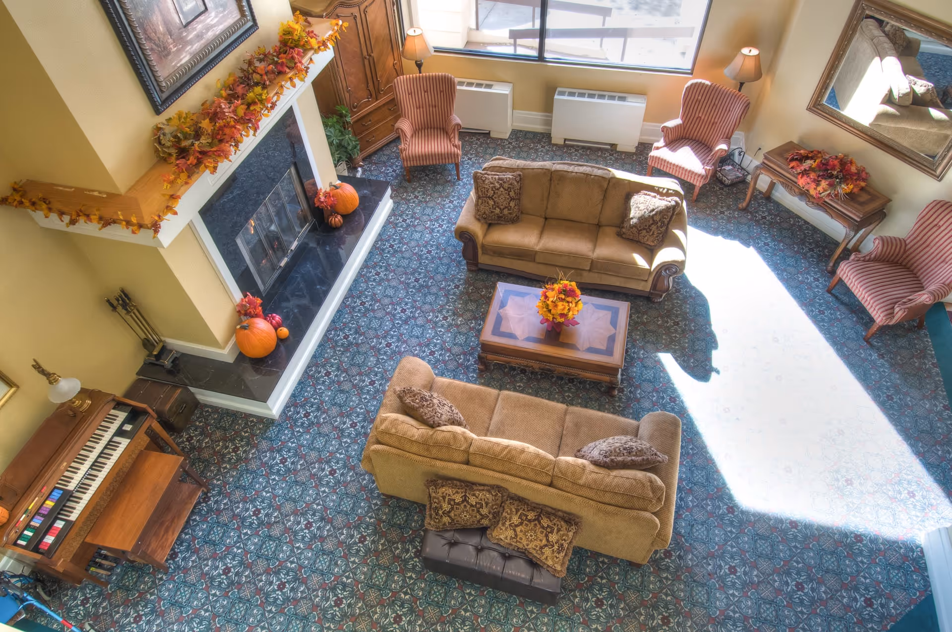 A cozy living room with two beige sofas, a wooden coffee table with a floral centerpiece, three striped armchairs, a fireplace decorated with autumn leaves and pumpkins, a piano, and a large window letting in sunlight.