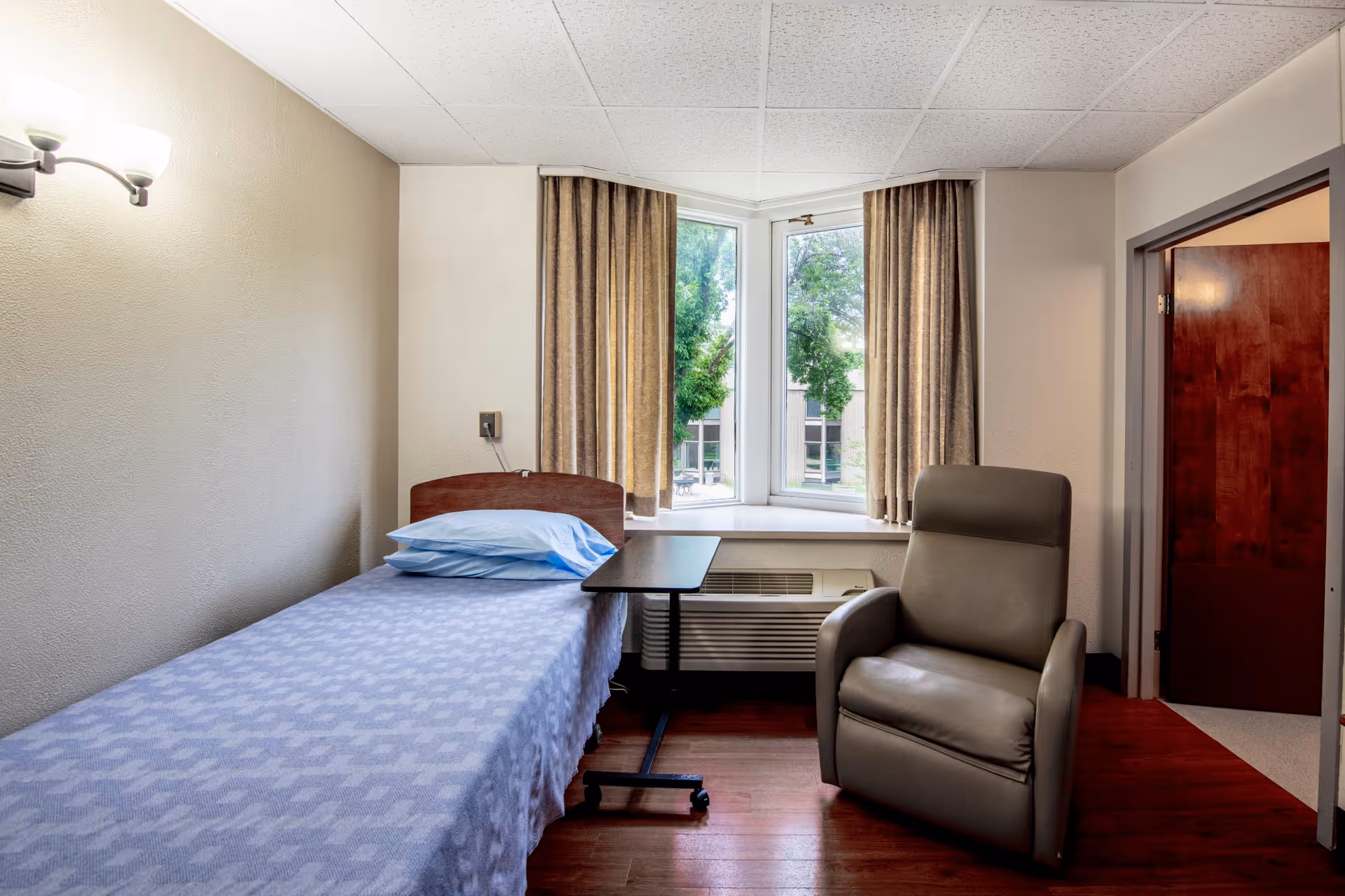 A small, simple bedroom in a senior living facility with a single bed covered in a blue patterned blanket and a blue pillow. Next to the bed is a small adjustable overbed table. A comfortable gray recliner chair is positioned near a window with beige curtains, and there is an air conditioning unit below the window. The room has light-colored walls and a wooden floor.