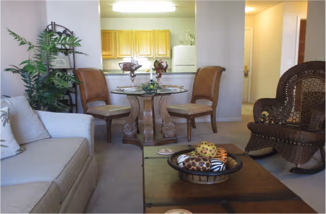 A cozy living room area with a beige sofa on the left, a wooden coffee table with decorative balls in a bowl, a wicker rocking chair on the right, and a small round dining table with two chairs in the background near a kitchen with wooden cabinets and a white refrigerator. There is also a green potted plant and a decorative shelf on the left side.