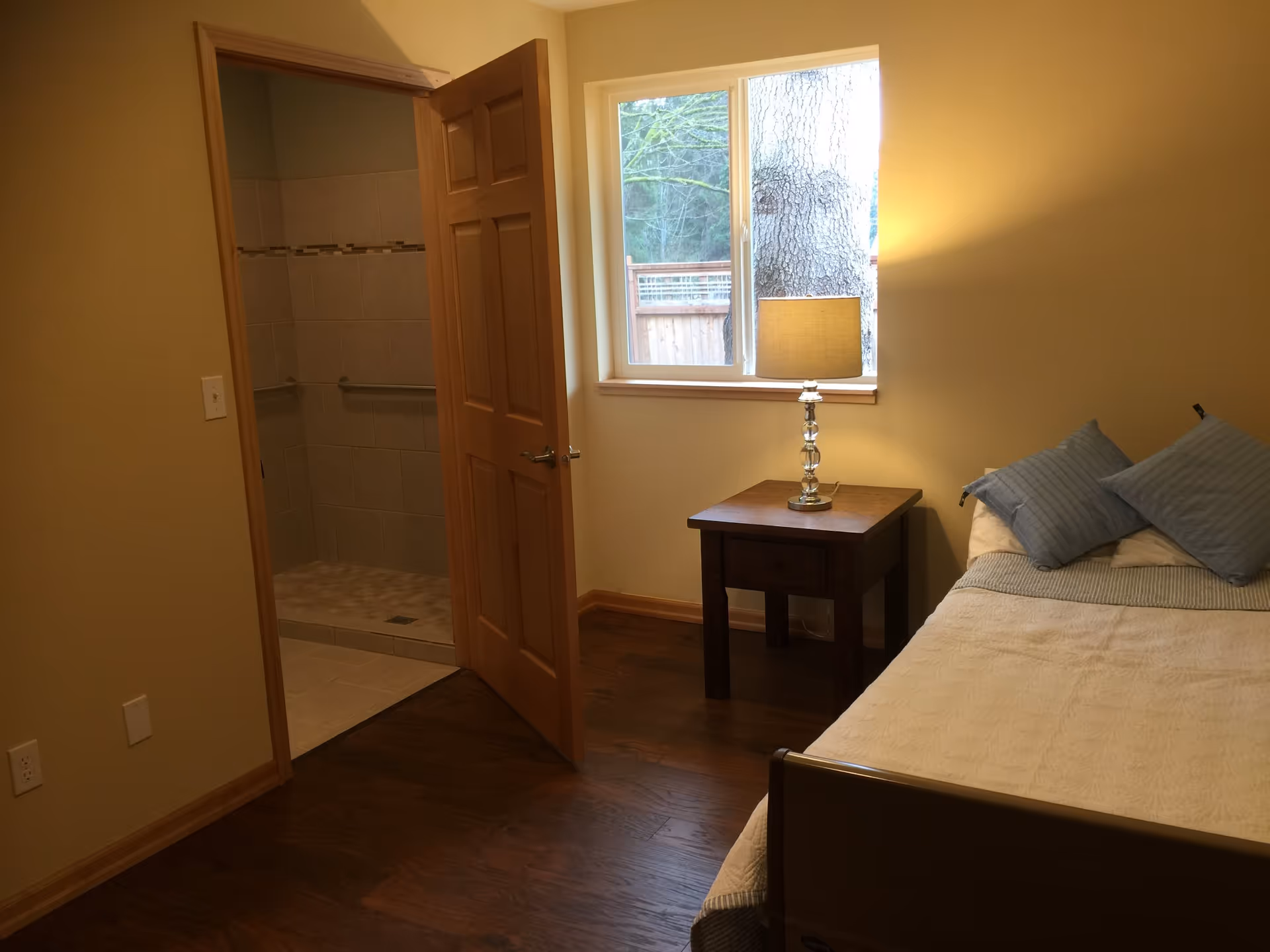 A bedroom with a wooden floor, a bed with white bedding and blue pillows, a wooden nightstand with a lamp, and an open door leading to a tiled bathroom with grab bars. A window next to the nightstand shows a tree and a wooden fence outside.