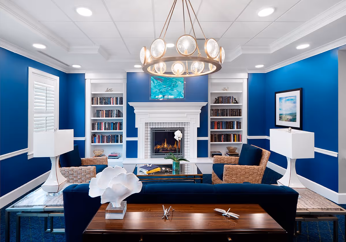 A cozy living room with bright blue walls, white trim, and a white fireplace in the center. The fireplace is flanked by built-in bookshelves filled with books. There are two wicker armchairs with blue cushions, a blue sofa, and a glass coffee table with books and a green vase with a white orchid. Two white table lamps sit on side tables beside the armchairs. A modern circular chandelier hangs from the ceiling, and there is artwork above the fireplace and on the right wall.