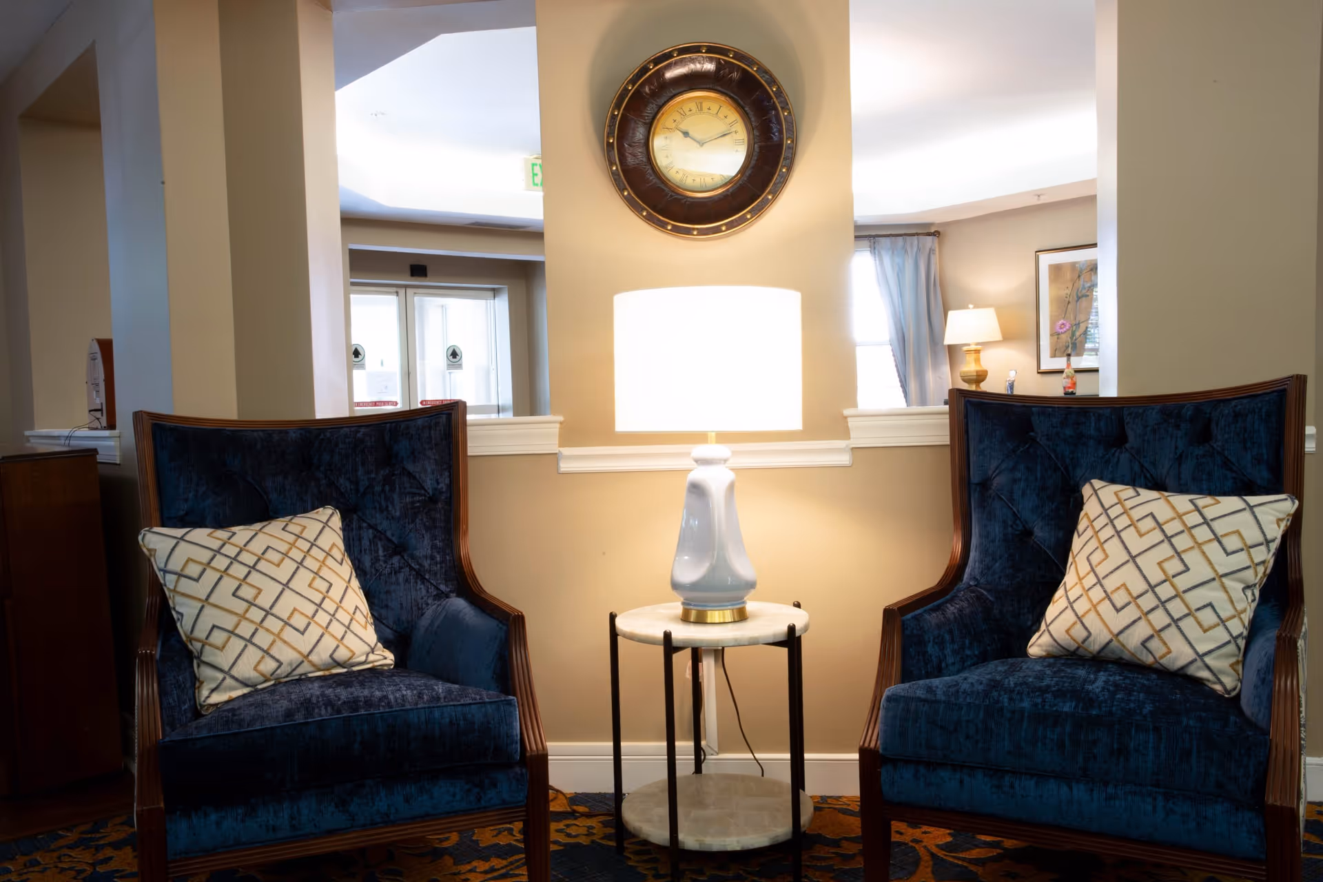 Two blue upholstered armchairs with patterned cushions flank a small round table with a white lamp on it. A round wall clock hangs above the lamp on a beige wall. The background shows part of a hallway and a room with a lamp and framed artwork.