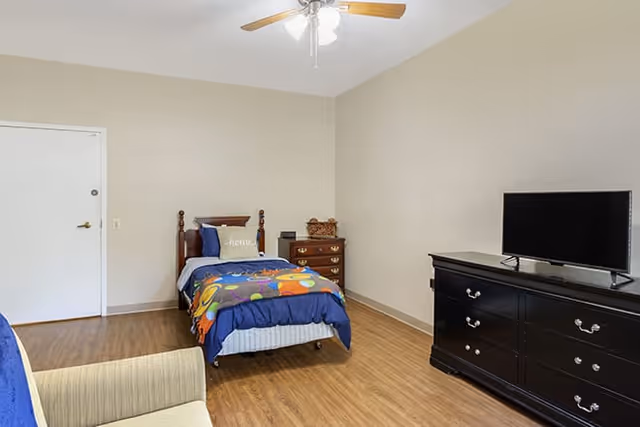 A simple bedroom in an assisted living community featuring a single bed with colorful bedding, a wooden dresser with a basket on top, a flat-screen TV on a black dresser, a beige armchair, and a ceiling fan with lights. The walls are painted light beige and the floor is wood.