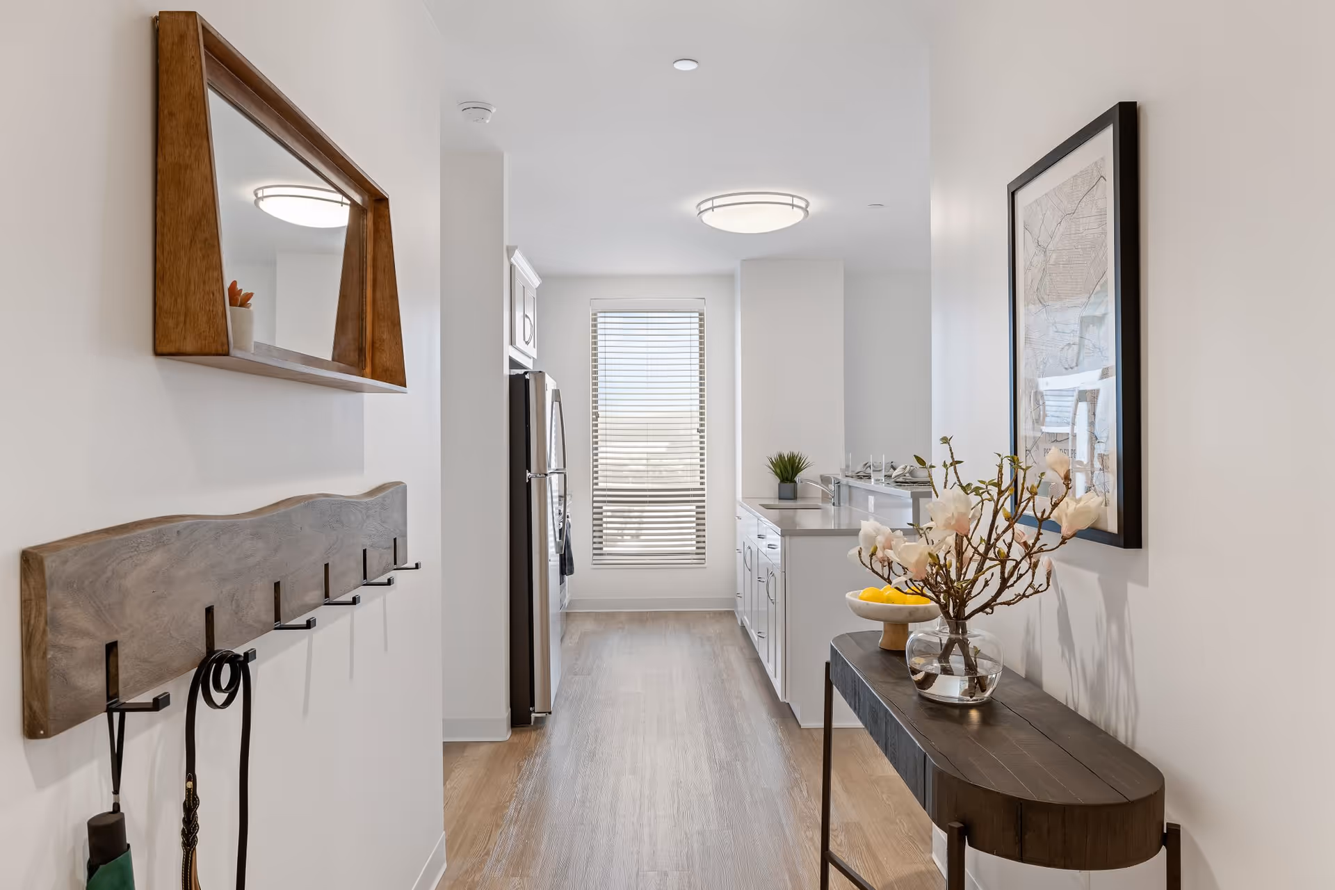 A bright, modern kitchen hallway with light wood flooring and white walls. On the left wall, there is a wooden coat rack with hooks and a wooden-framed mirror above it. At the end of the hallway, there is a window with blinds letting in natural light. On the right side, there is a narrow dark wood console table with a glass vase containing flowers and a bowl of lemons. A framed map or artwork hangs above the console table. The kitchen area features white cabinetry and stainless steel appliances.