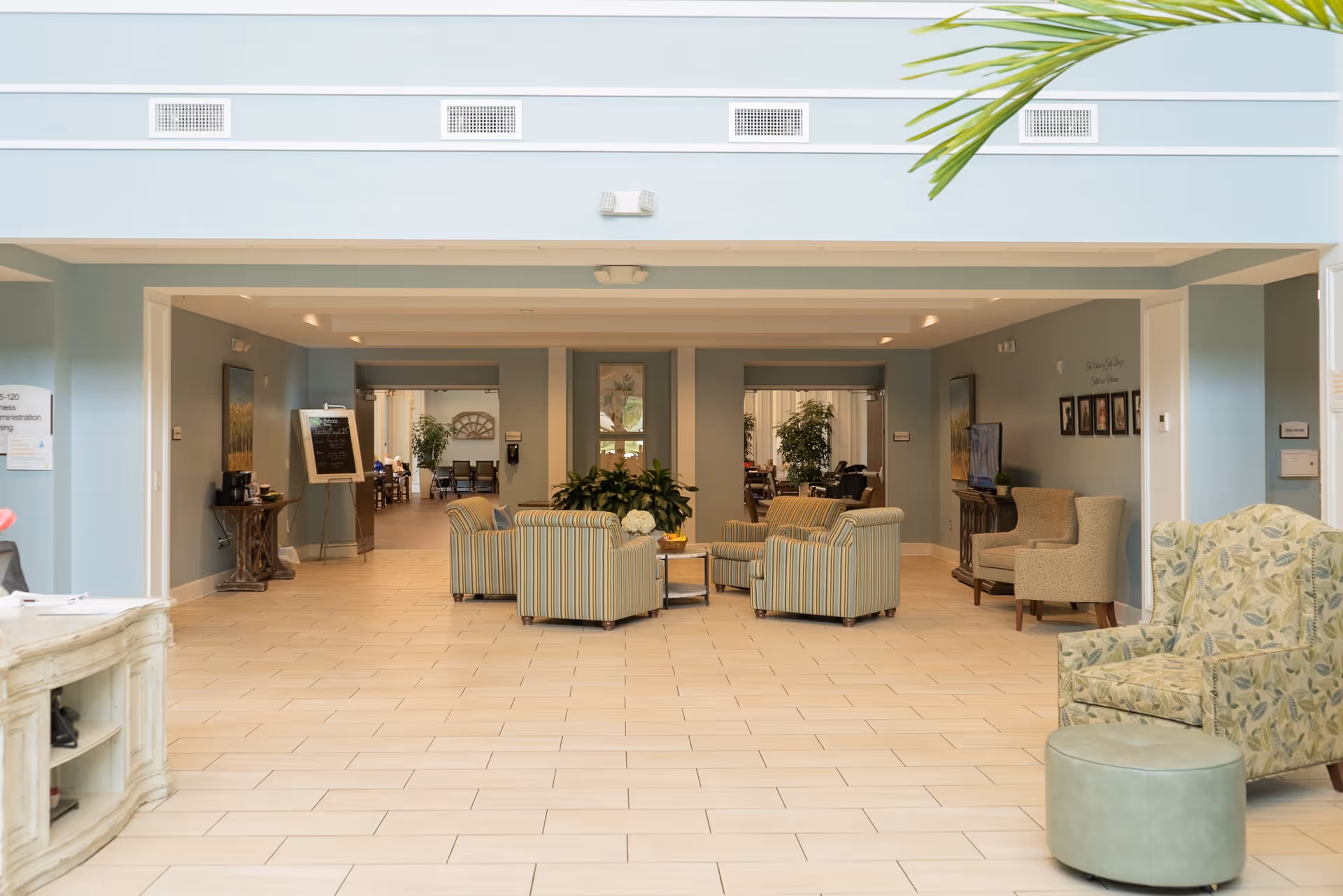 A spacious and well-lit common area with light blue walls and tiled floor. The room features several upholstered armchairs arranged around a small table with plants in the center. There are additional seating areas visible in the background, along with framed pictures on the walls and a TV on a stand. A green plant is partially visible in the upper right corner.