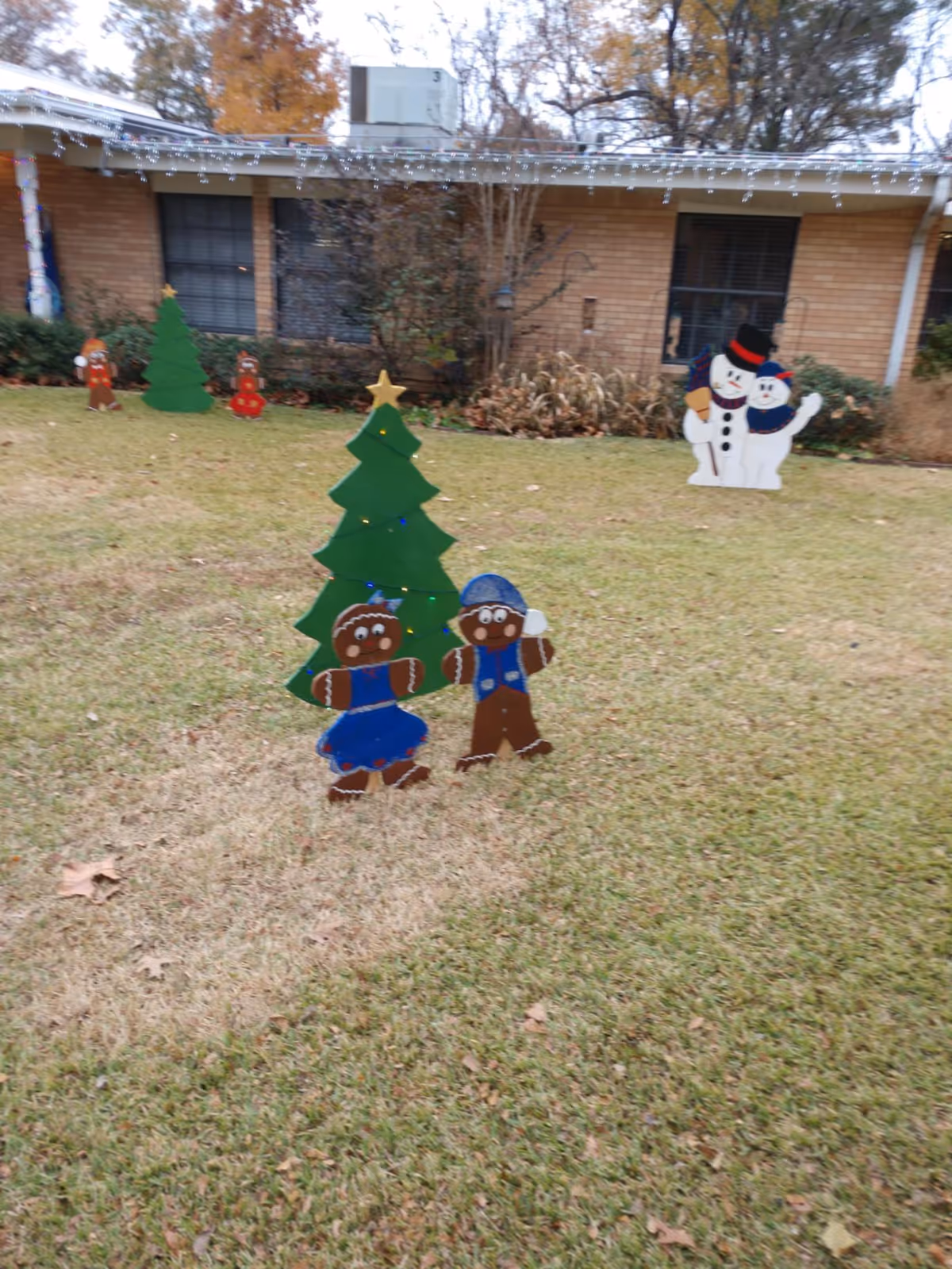 Outdoor lawn decorated with holiday-themed cutouts including gingerbread figures, Christmas trees, and snowmen in front of a brick building with windows and some bushes.