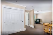 Interior view of a living space in an assisted living facility showing a hallway with white double closet doors on the left, a beige wall with a doorway leading to another room, a green armchair near a window, and a wooden dining table with chairs on the right.