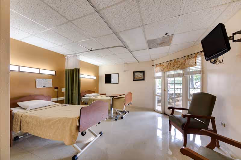 Shared nursing facility room with two hospital beds, chairs, a wall-mounted TV, and glass doors leading outside.