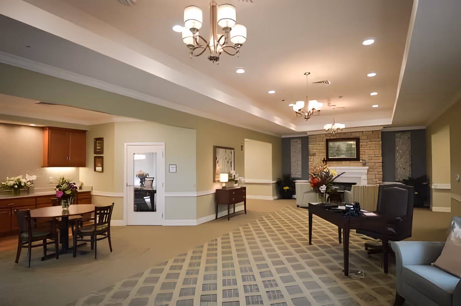 A spacious and well-lit common area in a senior living facility featuring a sitting area with armchairs and a table, a fireplace with a stone surround and a framed picture above it, a dining table with chairs near a kitchenette with wooden cabinets, and decorative lighting fixtures hanging from the ceiling.