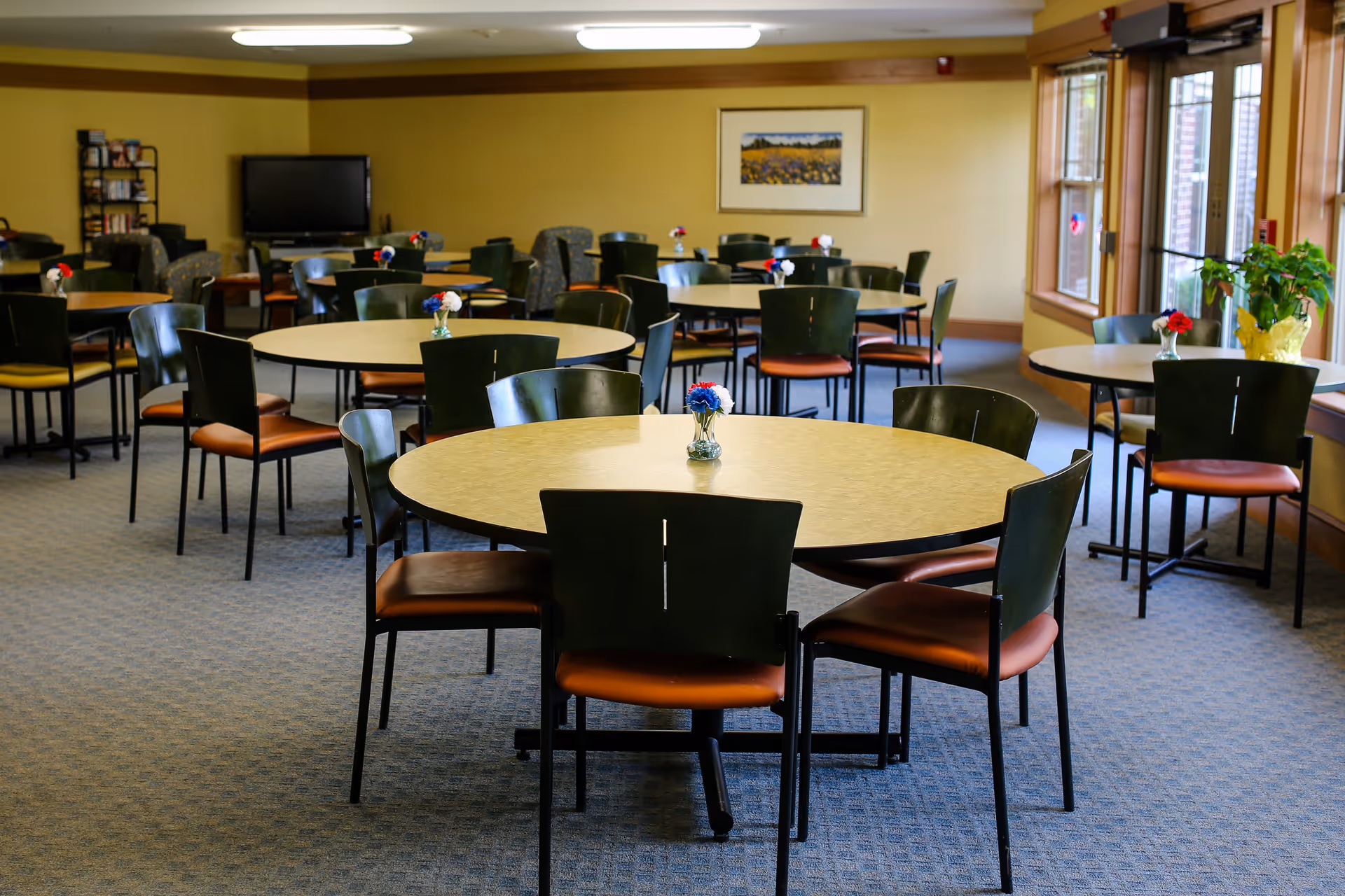 A spacious dining room with multiple round tables and black chairs with brown seats. Each table has a small vase with red, white, and blue flowers. The room has large windows on the right side letting in natural light, a carpeted floor, and a TV and bookshelf in the back corner.