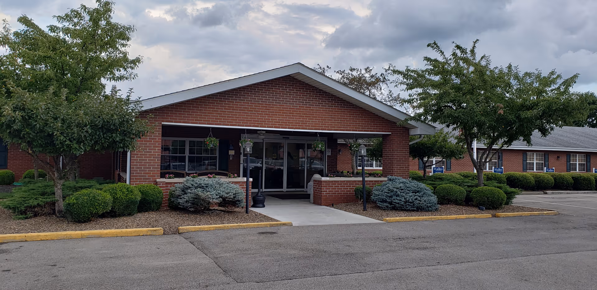 Front exterior view of a single-story brick building with a covered entrance, surrounded by neatly trimmed bushes and trees under a cloudy sky.