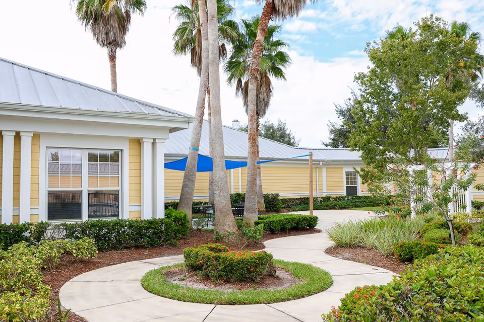 Outdoor courtyard area at The Brennity at Tradition Senior Living featuring a curved concrete walkway around a small landscaped island with palm trees, surrounded by bushes and greenery. Yellow buildings with white trim and metal roofs are visible in the background under a partly cloudy sky.