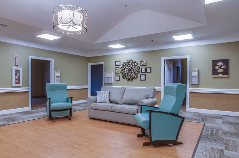 A common area in Barton House Memory Care featuring a gray sofa with a decorative pillow and two teal armchairs arranged on a wooden floor section surrounded by carpet. The walls are painted light green with a beige wainscoting and decorated with framed pictures and a decorative metal wall art. There are three doorways visible, ceiling lights, and a modern hanging light fixture.