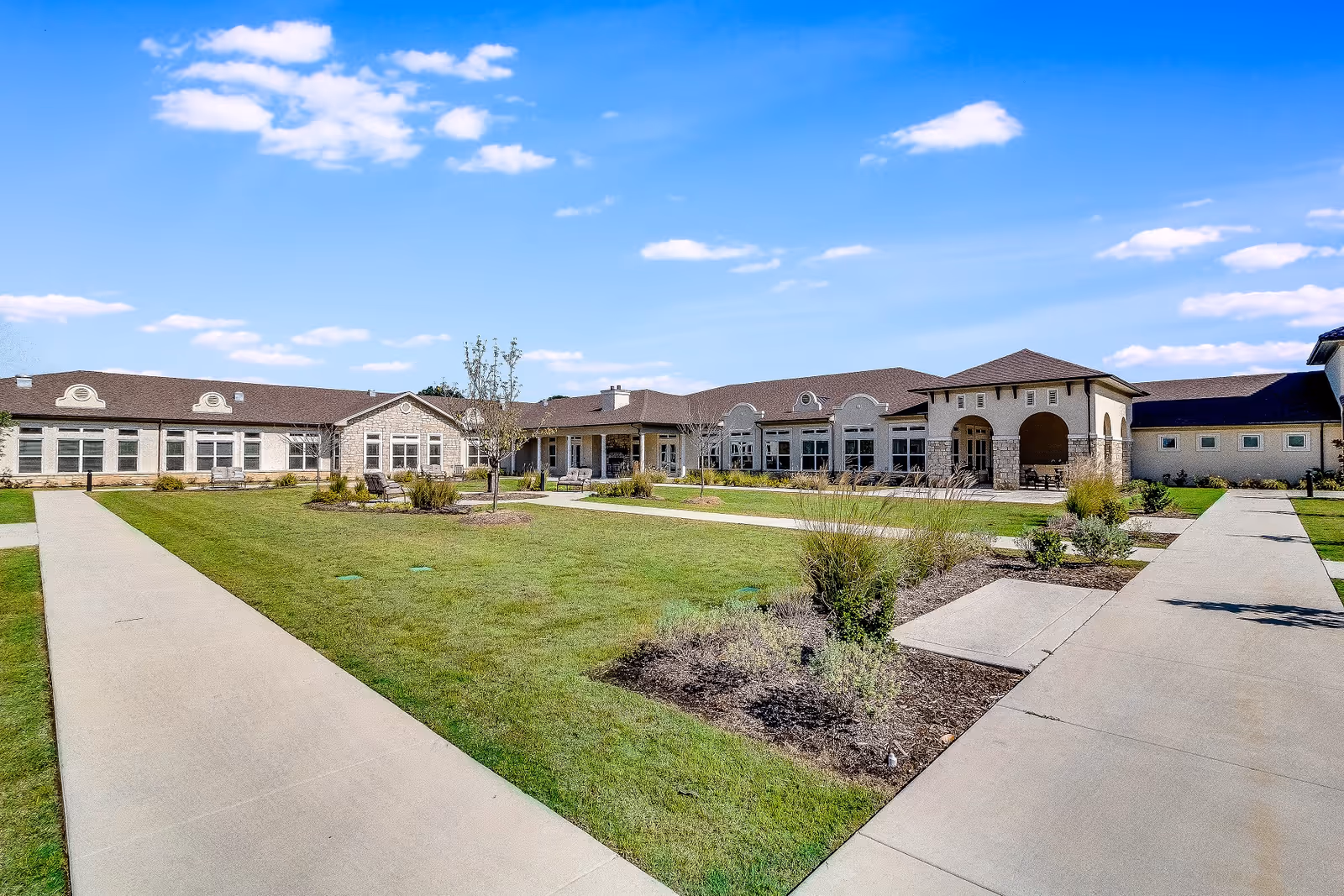 Courtyard with paved walkways and landscaping in front of a single-story senior living building under a blue sky.