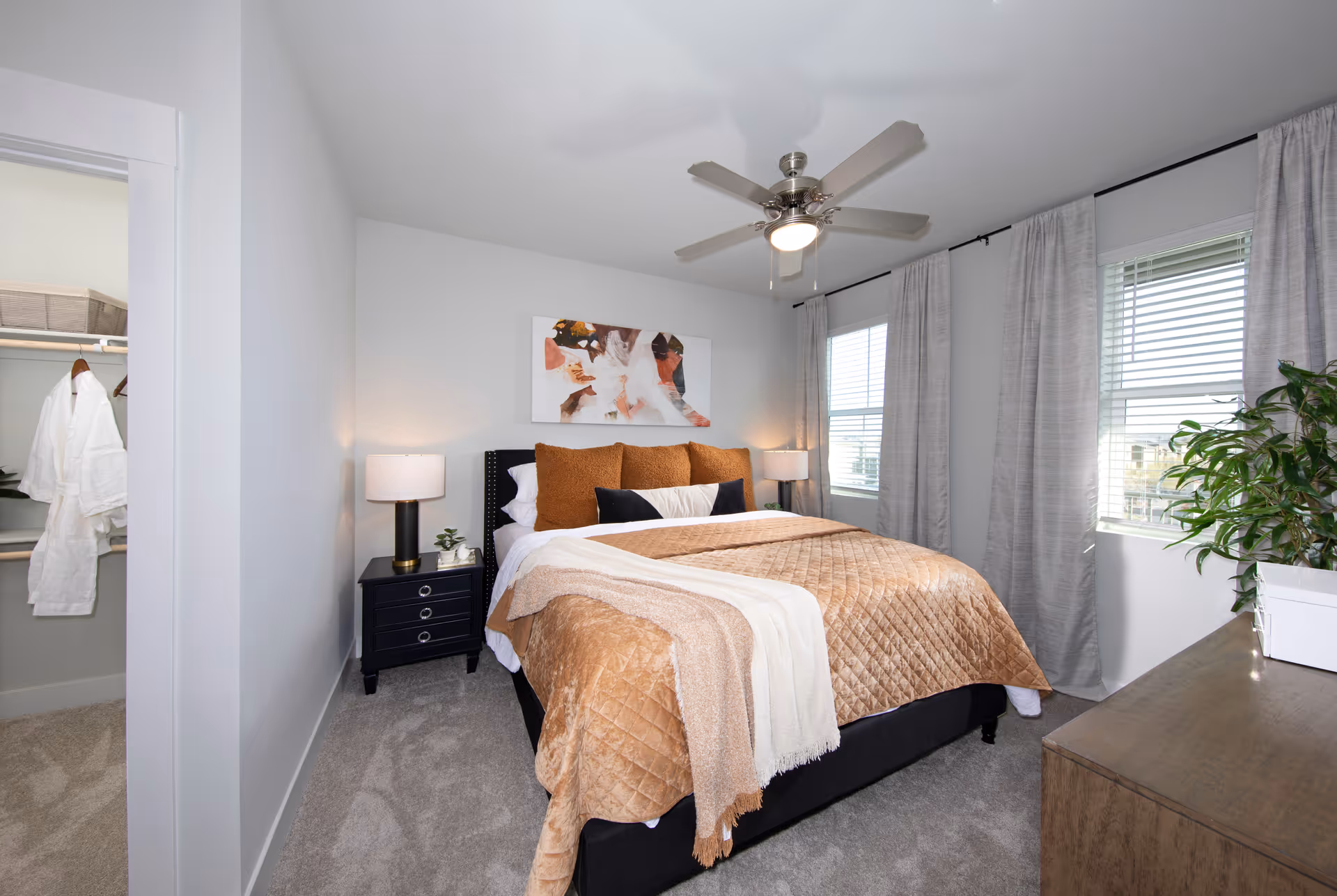 A neatly made bed with a tan quilt and multiple pillows in a bright bedroom. The room features two windows with gray curtains, a ceiling fan with a light, a black nightstand with a lamp and small plant, a wooden dresser with a plant on top, and an open closet with hanging robes.