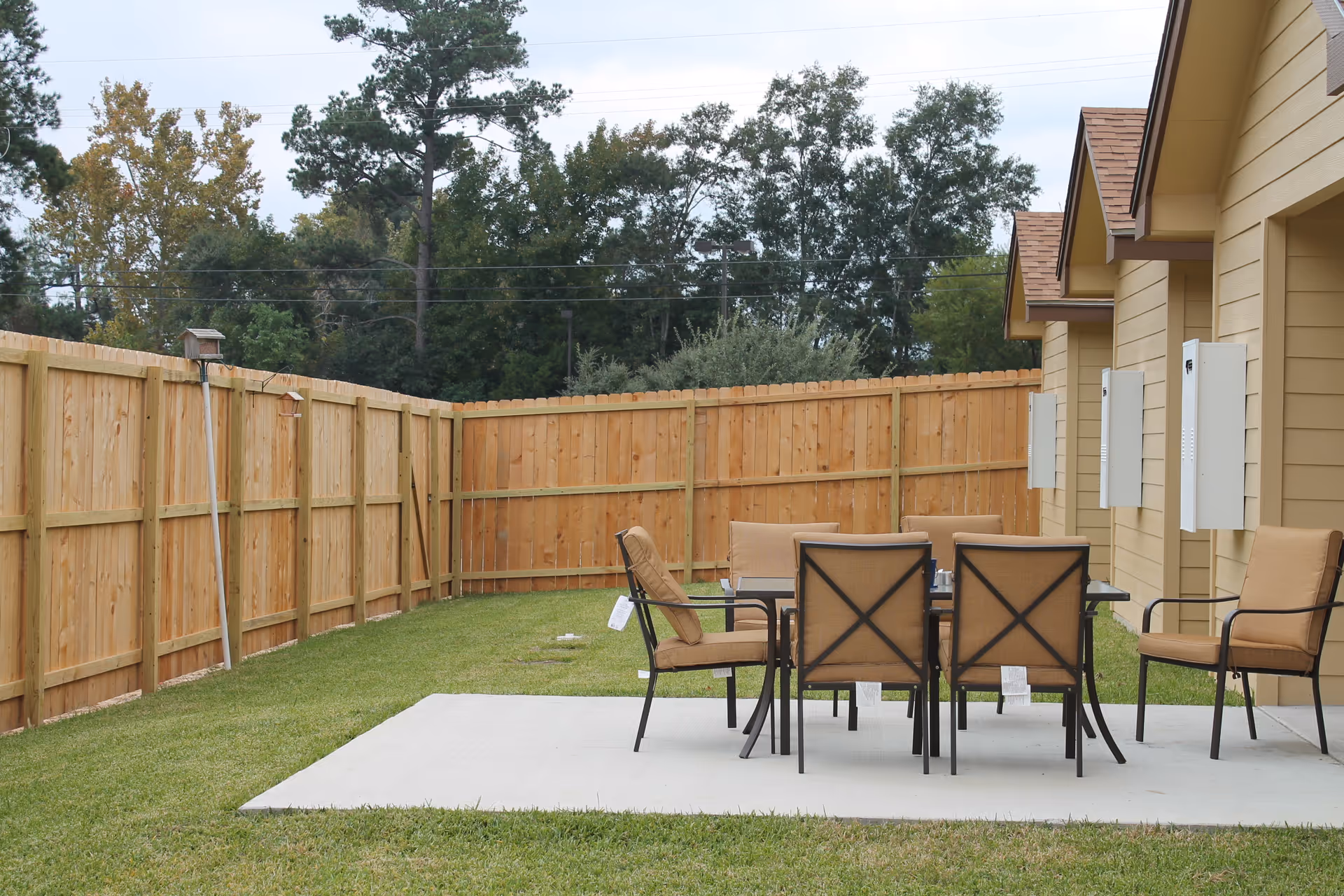 Outdoor patio area with a rectangular glass table surrounded by six cushioned chairs on a concrete slab, adjacent to a beige building with multiple doors and a wooden privacy fence enclosing the grassy yard.