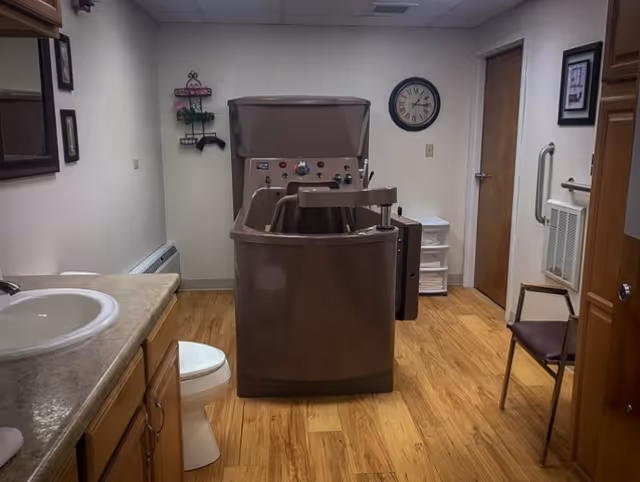 Interior room with a large brown mechanical bath device in the center, a white toilet and sink with countertop on the left, a wooden chair on the right, a wall clock, and a door in the background.