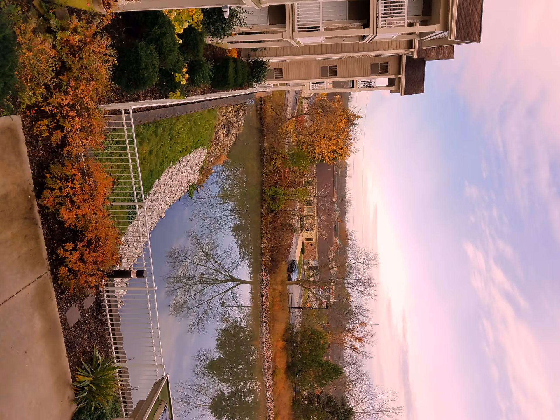 View of a landscaped outdoor area with a small pond or water feature surrounded by greenery and autumn-colored trees, adjacent to a multi-story residential building with balconies.