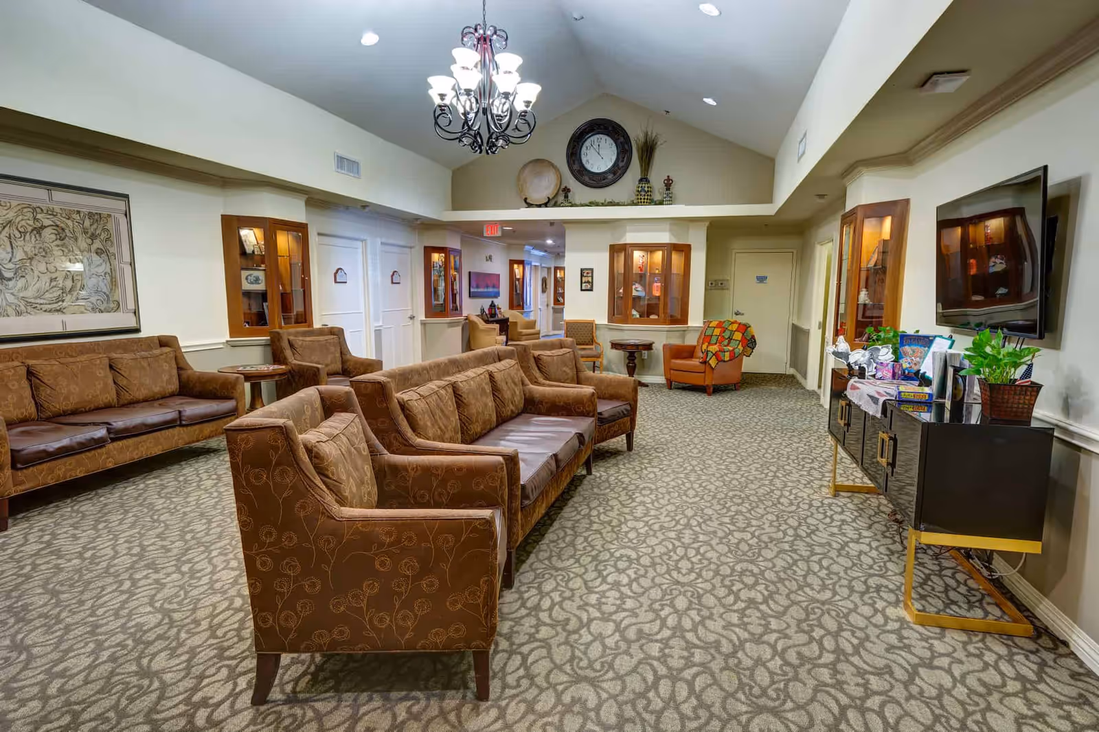 A furnished communal living room with brown sofas and armchairs, a wall-mounted TV, chandelier, and a large decorative clock above display shelves.