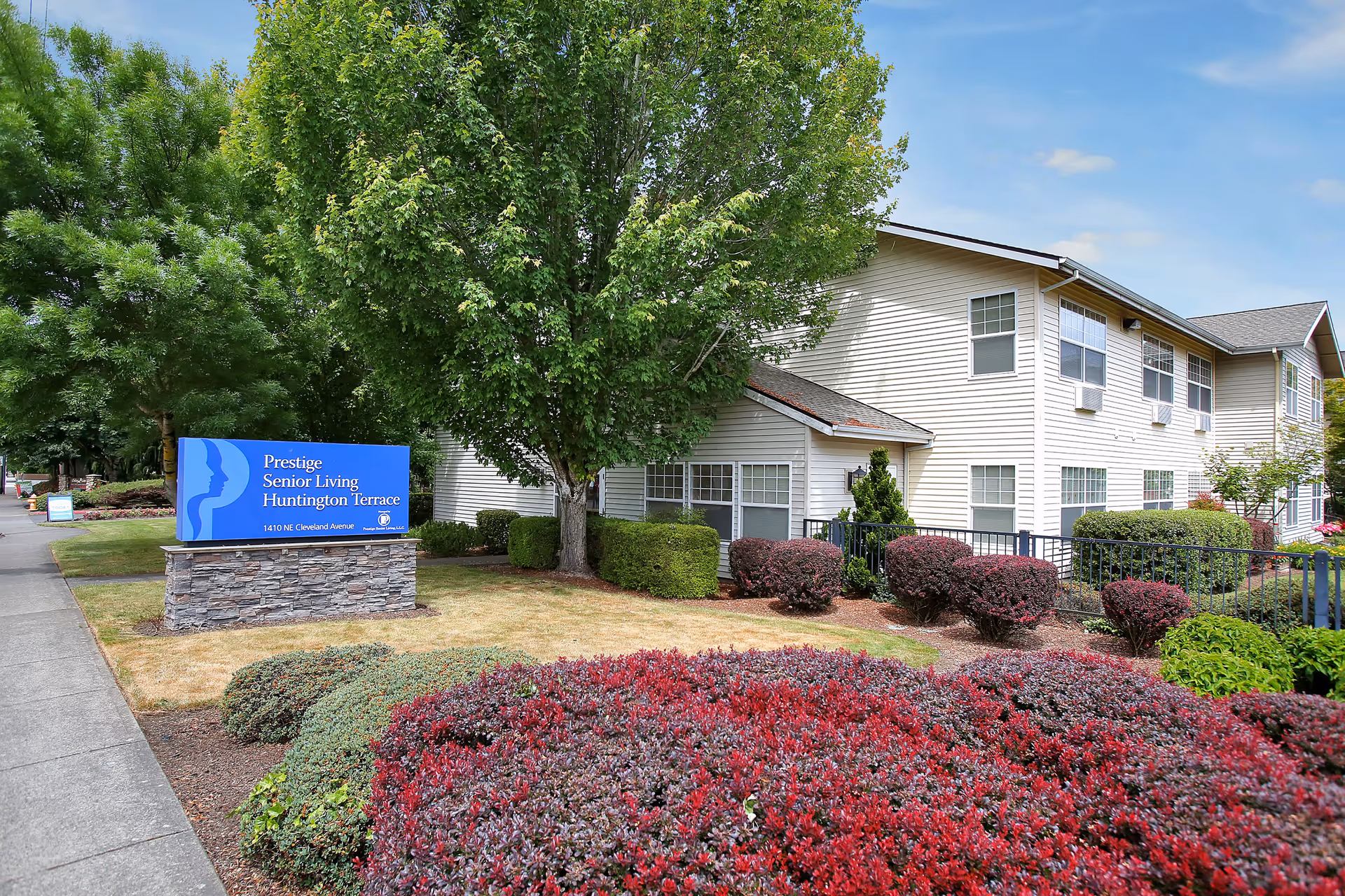 Exterior view of Prestige Senior Living Huntington Terrace building with well-maintained landscaping including bushes, trees, and red and green shrubs. A blue sign with the facility name and address is visible near the sidewalk.