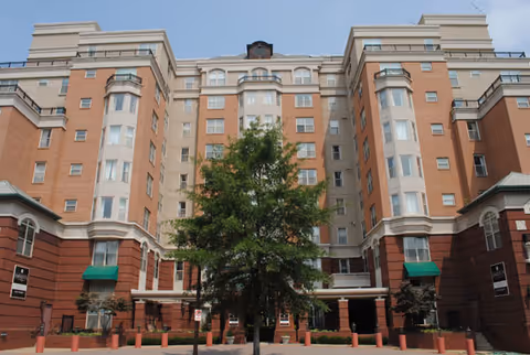 Front exterior of a multi-story brick senior living building with a central tree and covered entrance.