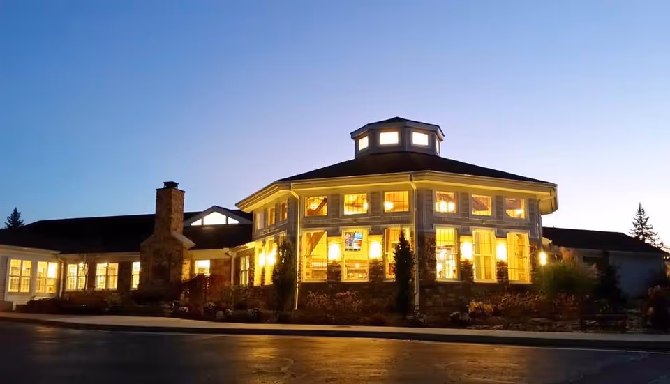 Exterior view of a well-lit retirement community building at dusk with large windows and a stone chimney, surrounded by landscaping and a paved driveway.