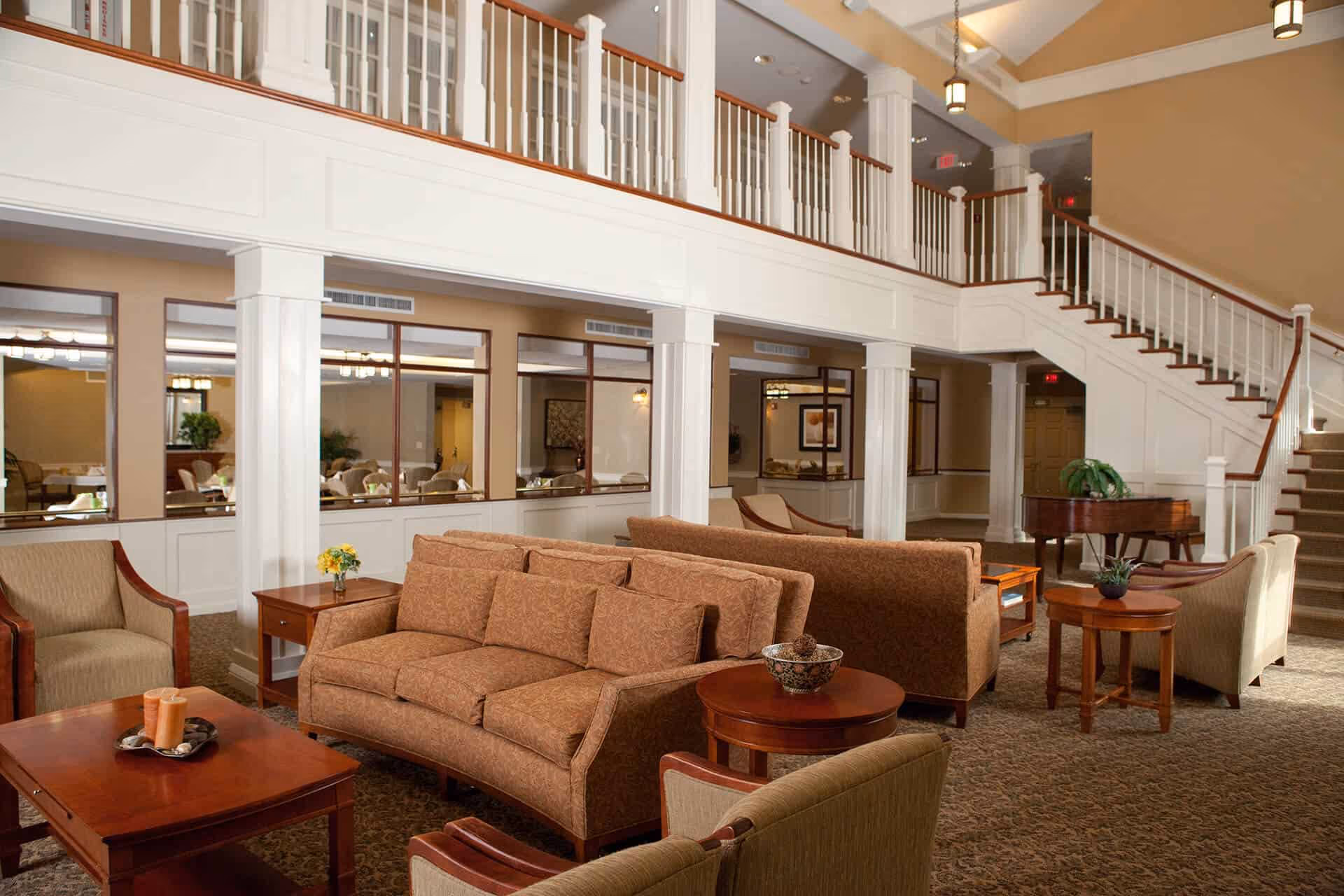A spacious and well-lit living room area in a senior living facility featuring comfortable upholstered sofas and armchairs arranged around wooden coffee and side tables. The room has a high ceiling with hanging light fixtures, white columns, and a staircase leading to an upper level with a white railing. There is a grand piano in the corner and large windows looking into an adjacent dining area.