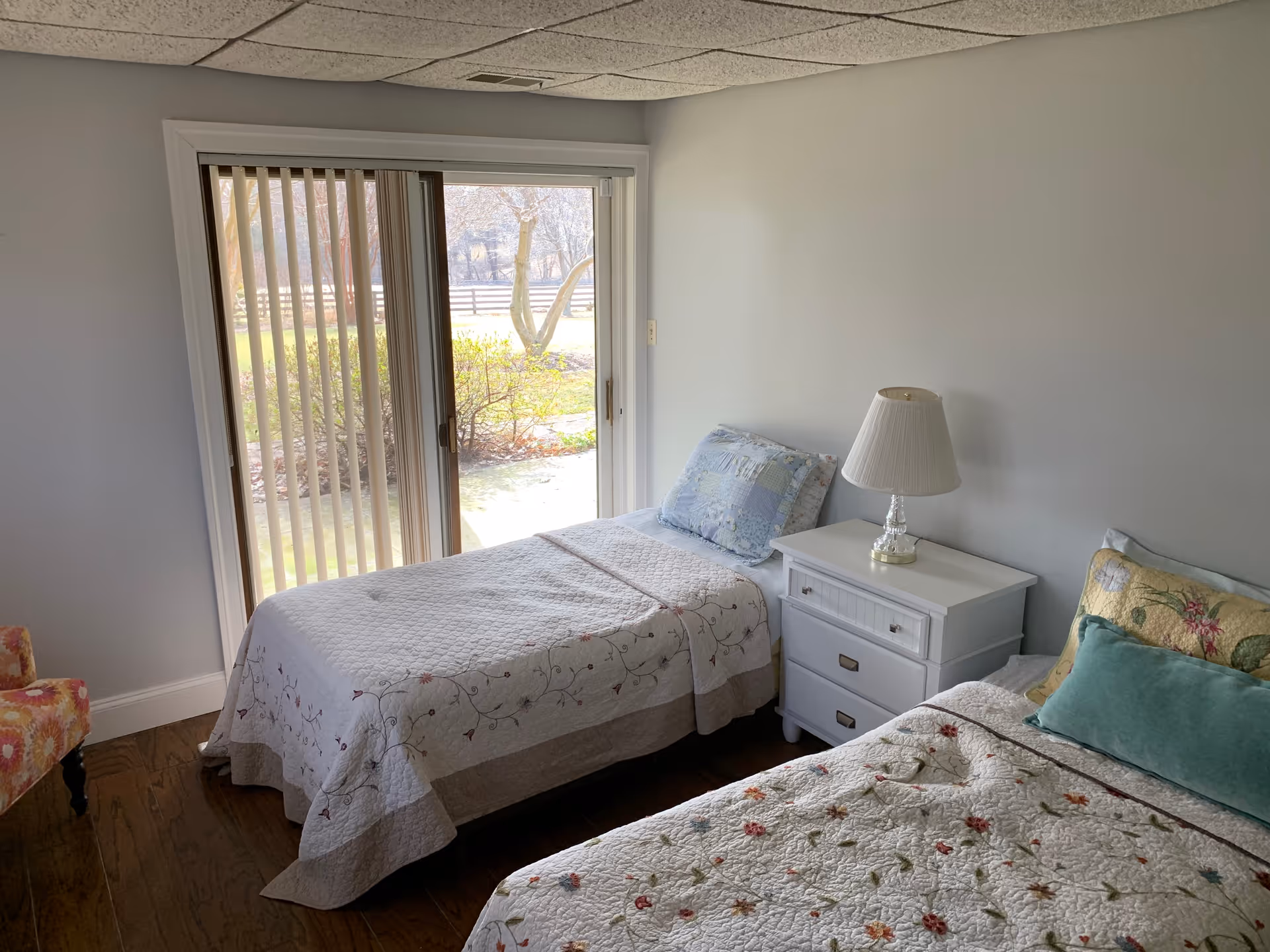 Bright bedroom with two twin beds, a white nightstand and lamp, and sliding glass doors with vertical blinds opening to a yard.