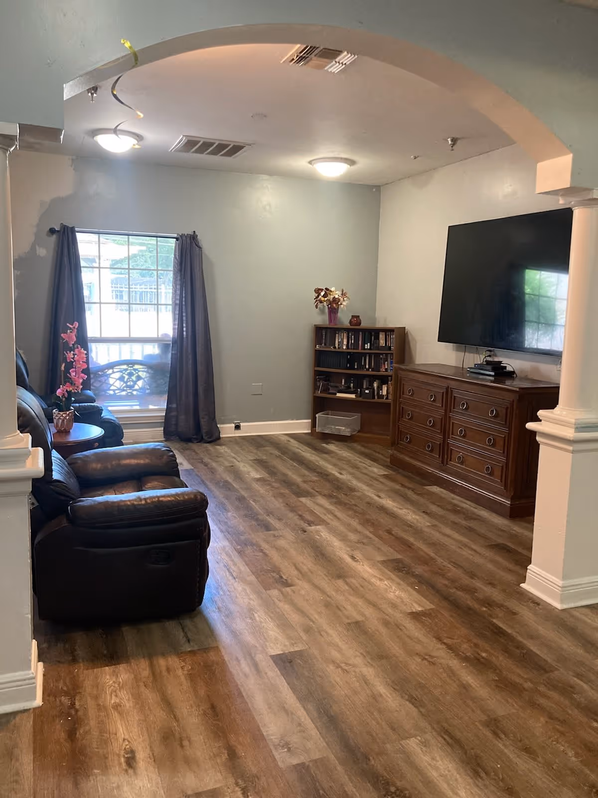 Living room with leather recliners, wood-look flooring, a wall-mounted TV above a dresser, a bookcase, and a window with curtains seen through an arched entry.
