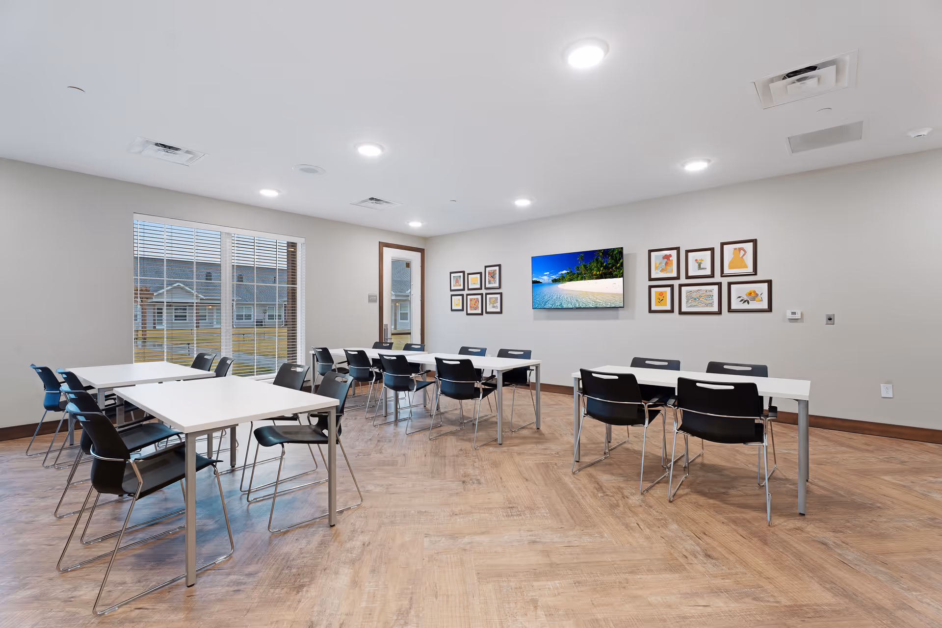 A bright, modern room with several white tables and black chairs arranged in small groups. The room has light wood flooring, a large window with blinds, and a door with a window. On the wall, there is a flat-screen TV displaying a beach scene and several framed pictures arranged around it.