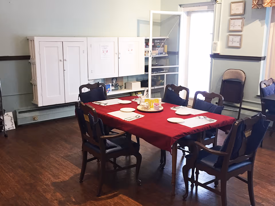 A communal dining room table covered with a red tablecloth set with plates and utensils, surrounded by wooden chairs and cabinets against pale green walls.
