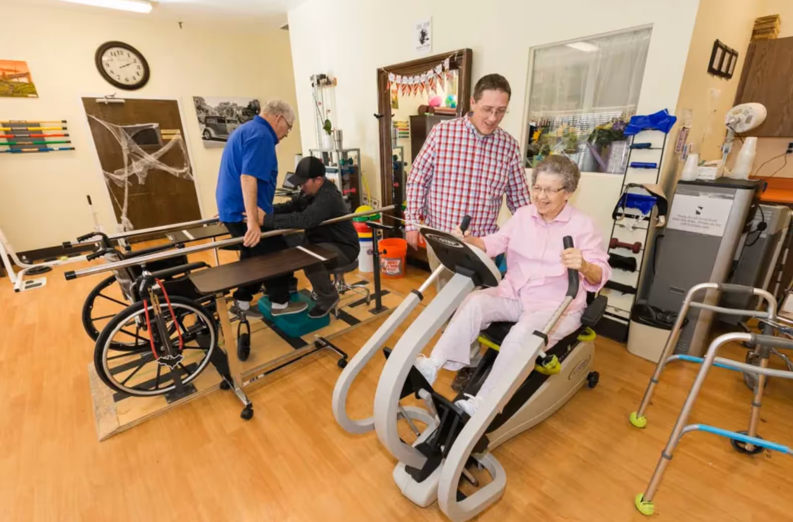 An elderly woman using a seated exercise machine in a rehabilitation center while a man in a checkered shirt assists her. In the background, another elderly man is receiving help from a caregiver with walking exercises using parallel bars. The room has wooden flooring and various rehabilitation equipment including walkers and wheelchairs.