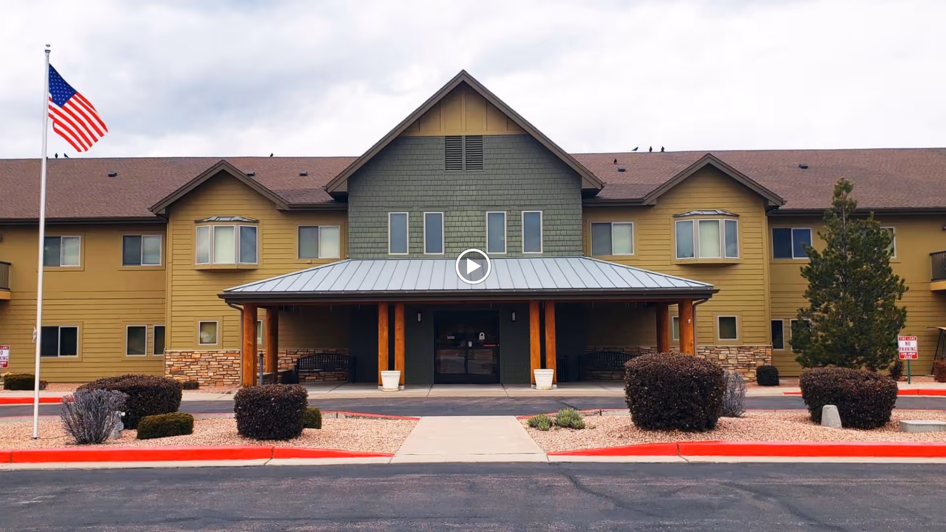 Front exterior view of a two-story retirement living facility building with a covered entrance supported by wooden pillars, landscaped bushes, and an American flag on a flagpole to the left.