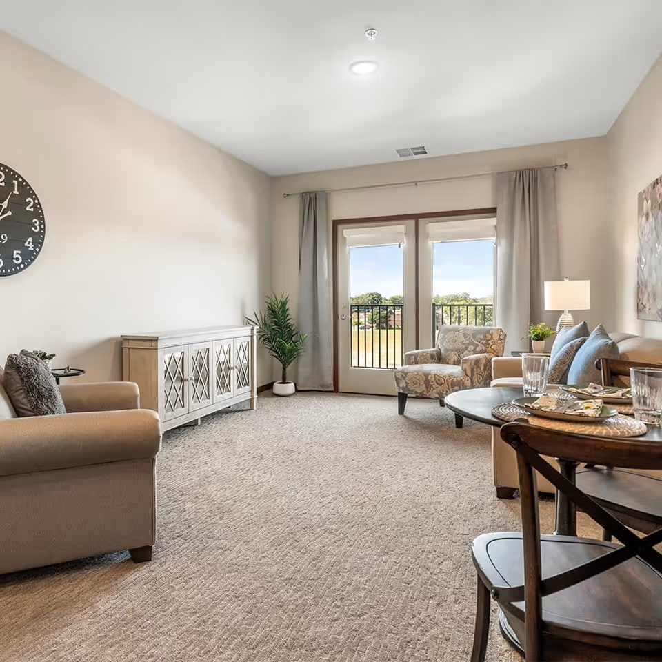 A bright and spacious living room in a senior living facility with beige walls and carpet. The room features a beige armchair, a patterned armchair, a wooden sideboard, a round dining table set with plates and glasses, a floor lamp, a potted plant, and large glass doors leading to a balcony with a view of greenery outside.