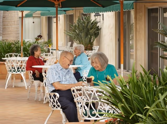 Four elderly people sitting at white wrought iron tables with green umbrellas on a tiled patio outside a building, enjoying drinks and conversation surrounded by plants.