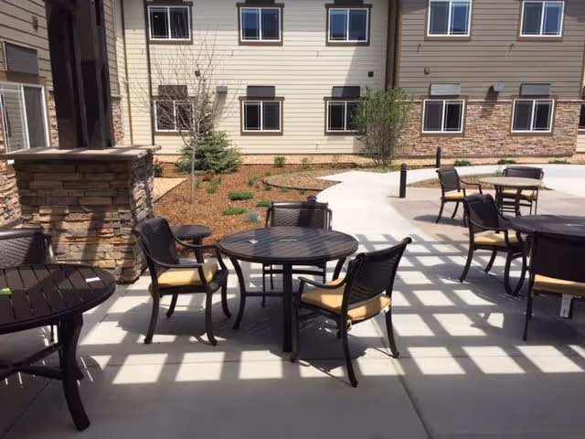 Outdoor patio area with several round tables and chairs with yellow cushions, adjacent to a building with beige siding and stone accents. There are small landscaped areas with mulch and young trees, and shadows from a pergola structure fall on the concrete floor.
