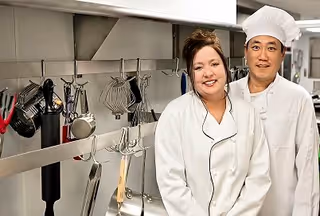 Two chefs in white uniforms standing side by side in a professional kitchen with various cooking utensils hanging on the wall behind them.