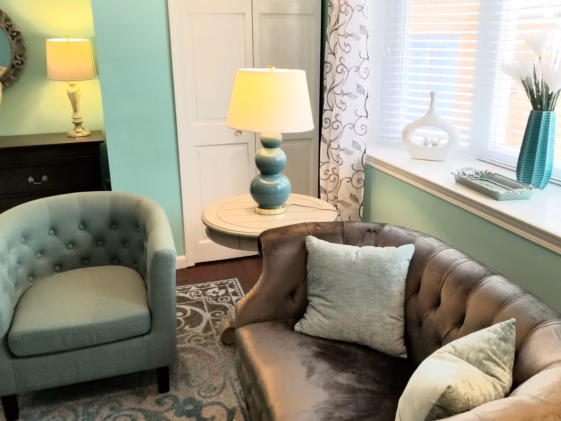 Cozy seating area with a tufted armchair and sofa, a side table with a lamp, and decorative items on a window ledge in a pastel-colored room.