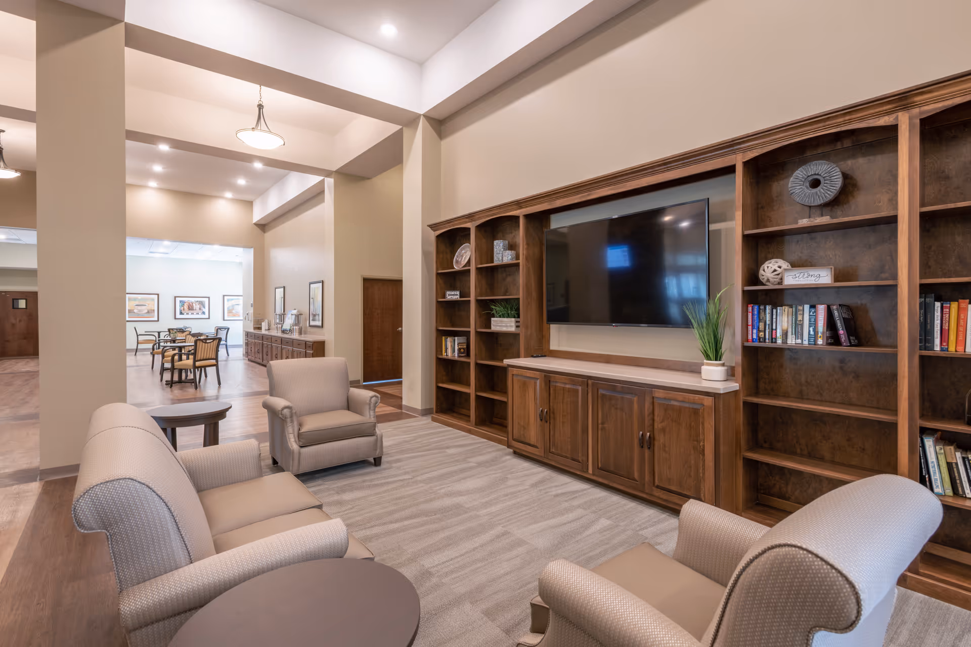 A cozy living room area in a senior living facility with beige upholstered armchairs and a sofa arranged around a round wooden coffee table. A large wooden entertainment center with built-in shelves holds a flat-screen TV, books, and decorative items. The room has soft lighting and a carpeted floor, with a dining area visible in the background.