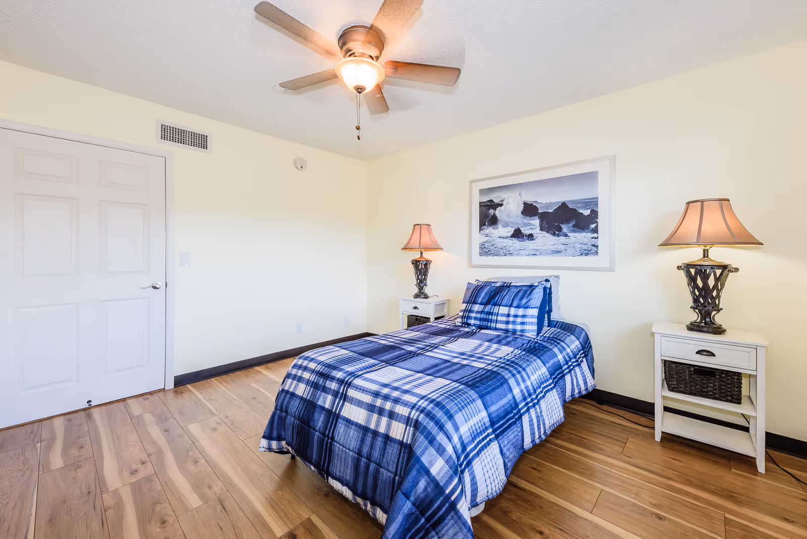 A bedroom with a single bed covered in a blue and white plaid comforter and matching pillow. There are two white nightstands on either side of the bed, each with a lamp. Above the bed hangs a framed picture of ocean waves crashing against rocks. The room has light yellow walls, wood flooring, a ceiling fan with a light, and a closed white door.