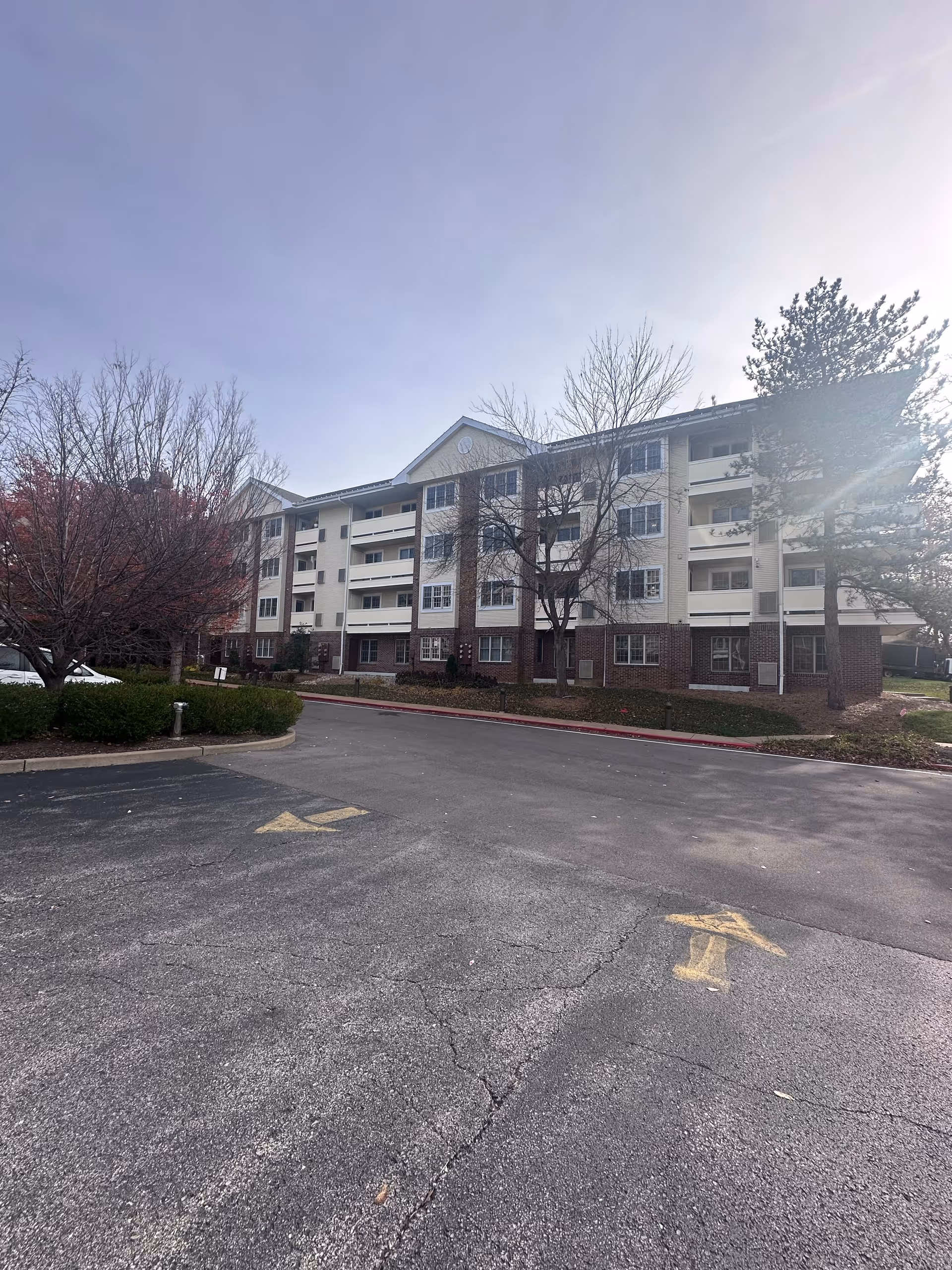 Four-story beige-and-brick senior living building with balconies overlooking a mostly empty parking lot and leafless trees.