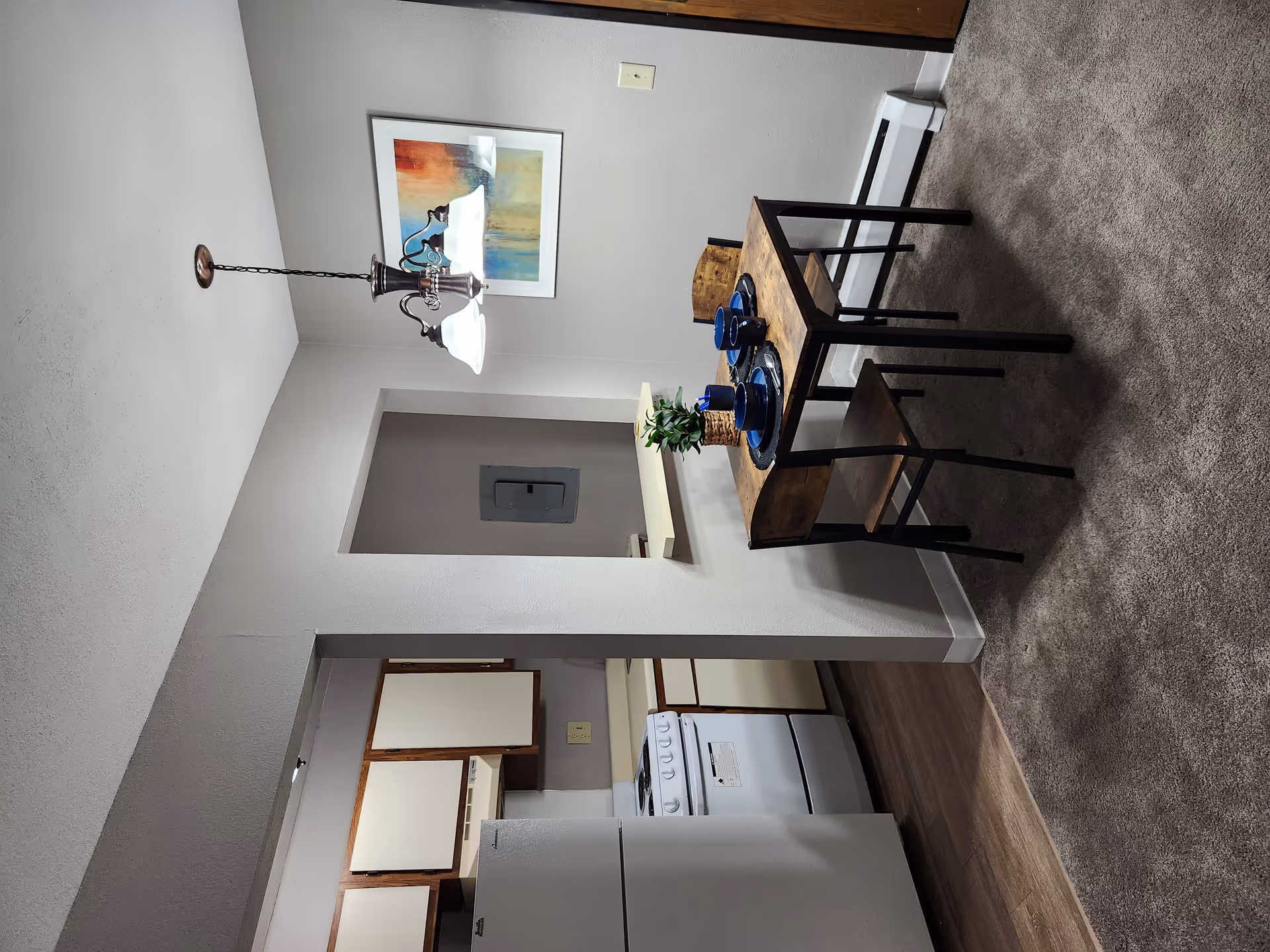 Interior view of a small dining area and kitchen in a senior living facility. The dining area has a wooden table with two matching chairs, set with blue plates and cups. A small potted plant is on the table. The kitchen features white appliances including a refrigerator and stove, with wooden cabinets. A hanging light fixture is above the dining table, and a colorful abstract painting is on the wall.