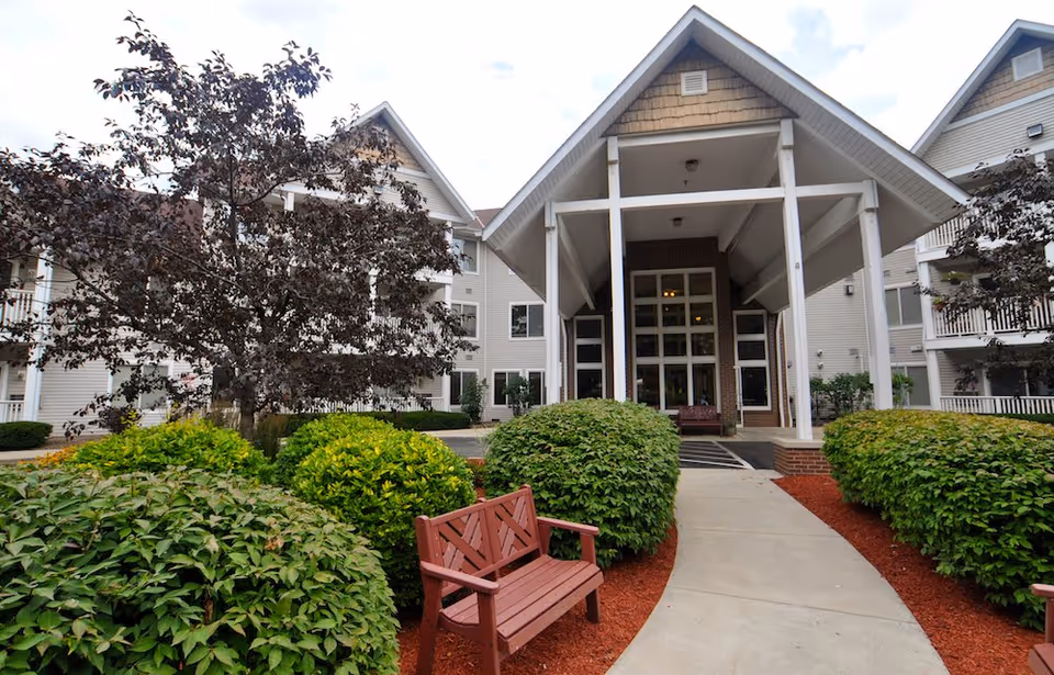 Entrance of a senior living facility with a covered porch supported by white pillars, surrounded by green bushes and trees. A wooden bench is placed along the curved concrete pathway leading to the entrance. The building is light-colored with multiple windows and balconies.