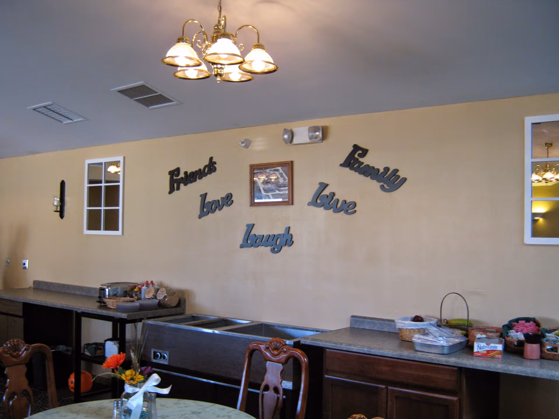 Interior view of a dining area with a countertop holding baskets and food items. The wall has decorative words saying 'Friends', 'Love', 'Family', 'Live', and 'Laugh'. There are wooden chairs and a table with a flower vase in the foreground, and a chandelier hanging from the ceiling.