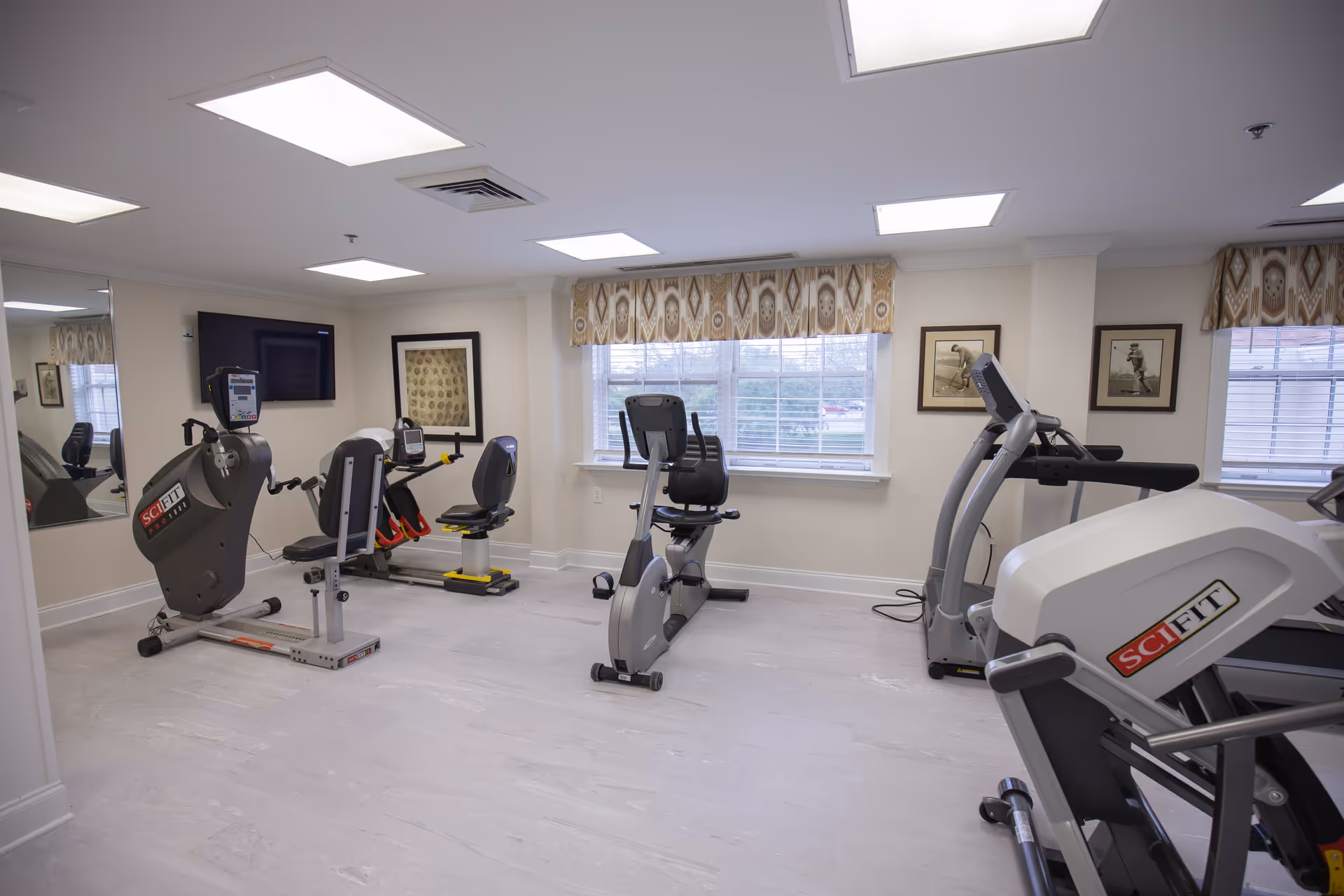Bright exercise room with several recumbent and upright cardio machines, a wall-mounted TV, and windows with patterned valances.