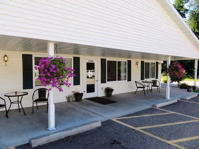 Exterior view of Care Partners Assisted Living-Stevens Point showing a white building with a covered porch. The porch has hanging baskets with purple flowers, small tables with chairs, and potted plants near the entrance door. There is a parking area with marked spaces in front of the porch.