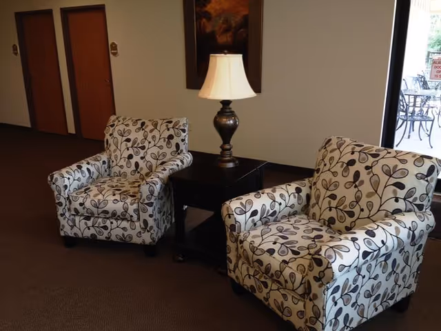 Two patterned armchairs with a leaf design are placed on either side of a dark wooden side table with a lamp on it. The setting appears to be an indoor common area with a window showing an outdoor patio with metal chairs and tables.
