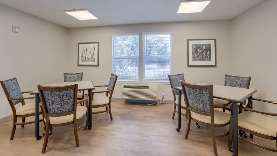A bright room with two rectangular tables, each surrounded by four wooden chairs with cushioned seats and patterned backs. The room has light-colored walls, two framed botanical prints on the walls, a large window with blinds, and a wall-mounted air conditioning unit below the window. The floor is wooden.