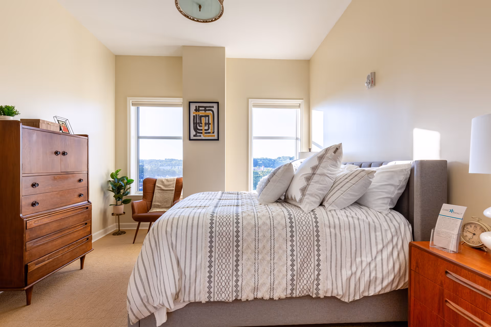 A bright bedroom with a neatly made bed featuring patterned white and gray bedding and multiple pillows. The room has beige walls and carpet, two windows letting in natural light, a wooden dresser on the left, a brown armchair with a throw blanket near the windows, a small plant, and a wooden nightstand with a lamp, clock, and informational pamphlet on the right.
